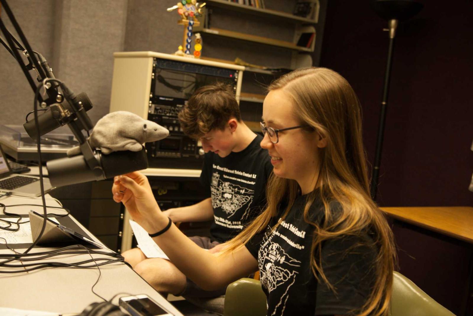 Students sitting in recording studio behind a microphone with a stuffed animal crouching on the mic.