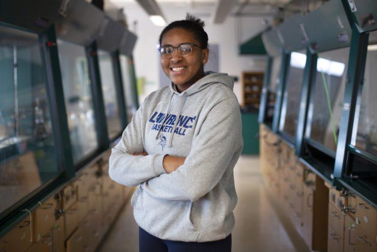 Kenya Earl, wearing a grey Lawrence basketball sweatshirt, stands with her arms crossed and smiles at the camera.