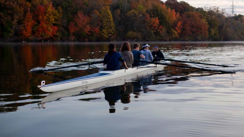Five students row down the Fox River against fall colored leaves in background.