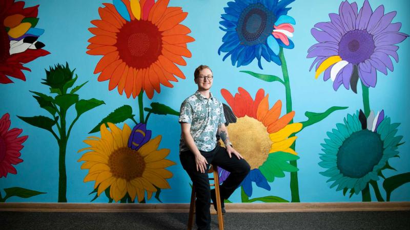 Cael Neary sits on a stool in front of his colorful mural in the Gender and Sexuality Diversity Center.