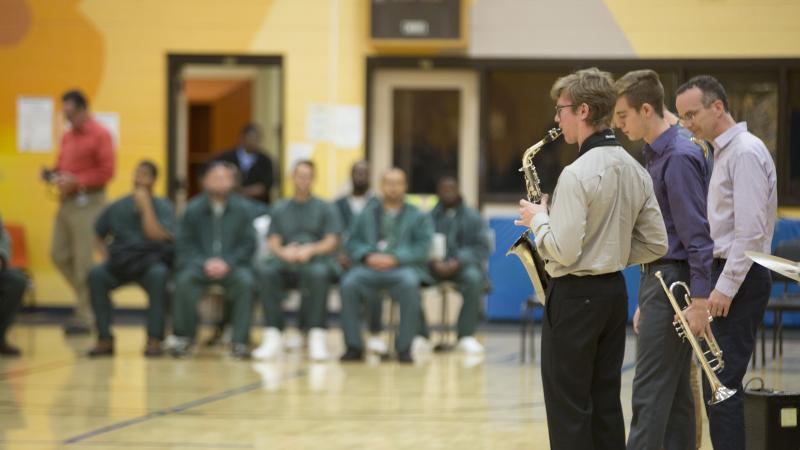 Jazz musicians play in a gynasium, with men in green jumpsuites in the background.
