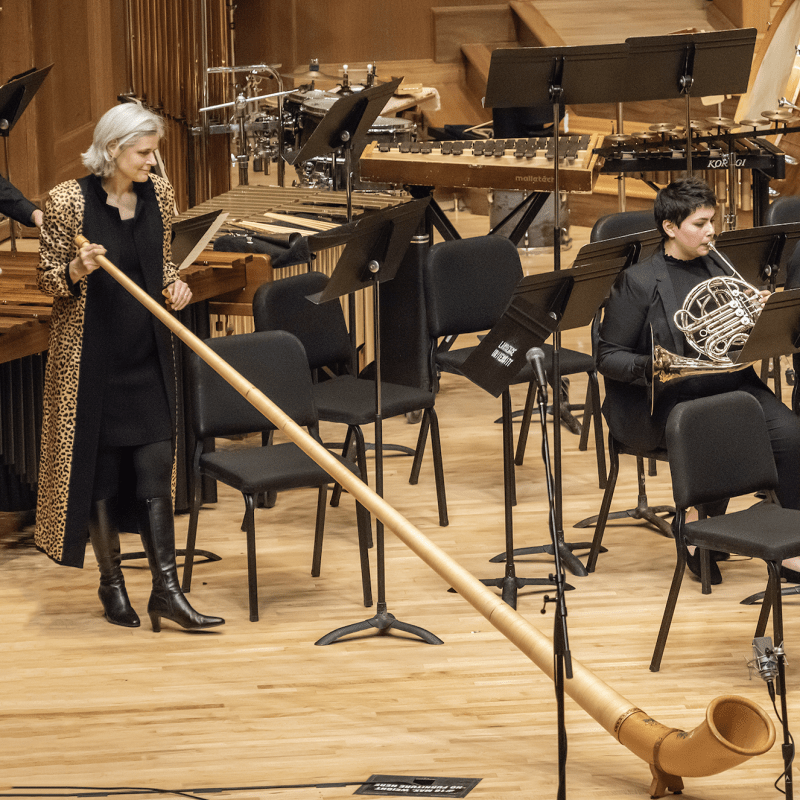 Woman with white hair holding an alphorn on stage with other musicians wearing a long coat with a leopard print