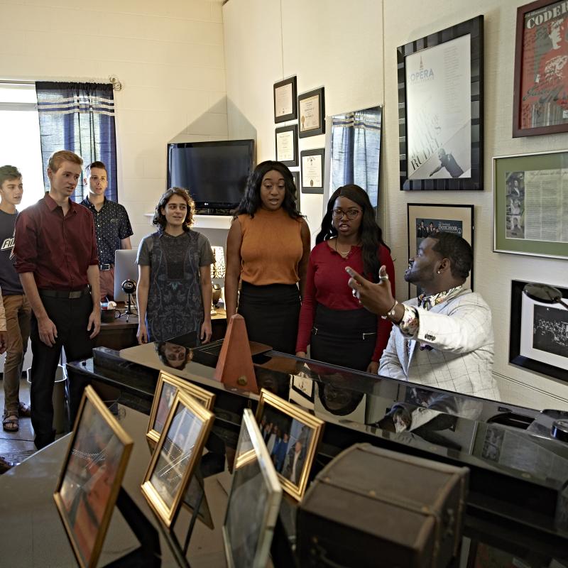 Students stand around a piano singing with a man playing