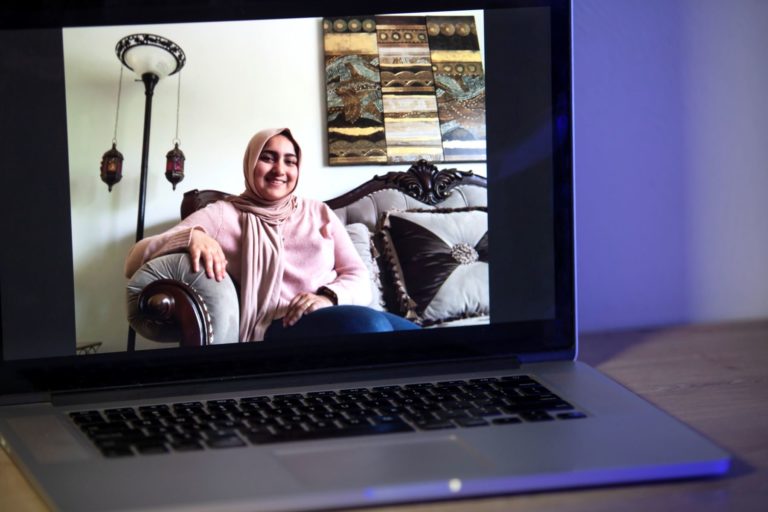 A laptop screen shows Ghania Imran, wearing a pink sweater and sitting on a grey couch.