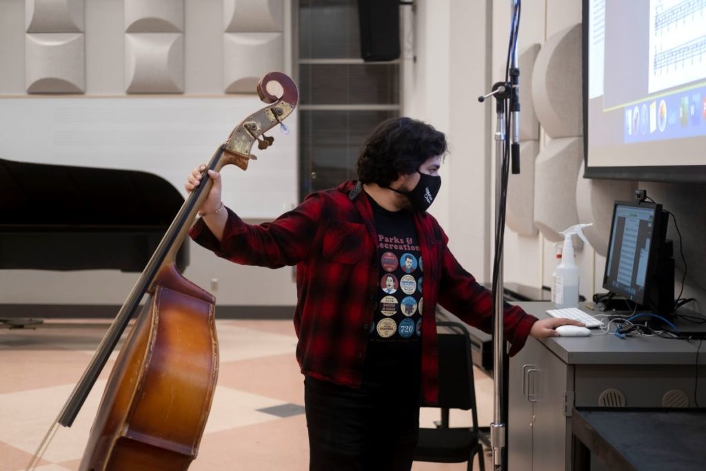 Student holding a double bass in one hand and working on a computer with the other