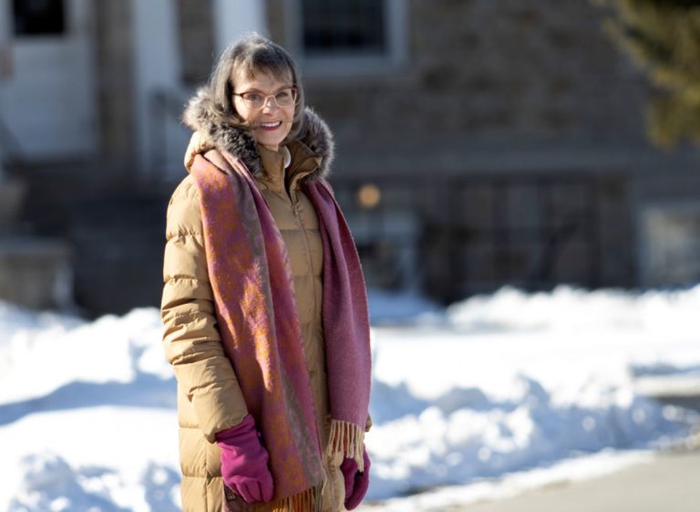 Eilene Hoft-March, wearing a tan coat and pink scarf, smiles at the camera.