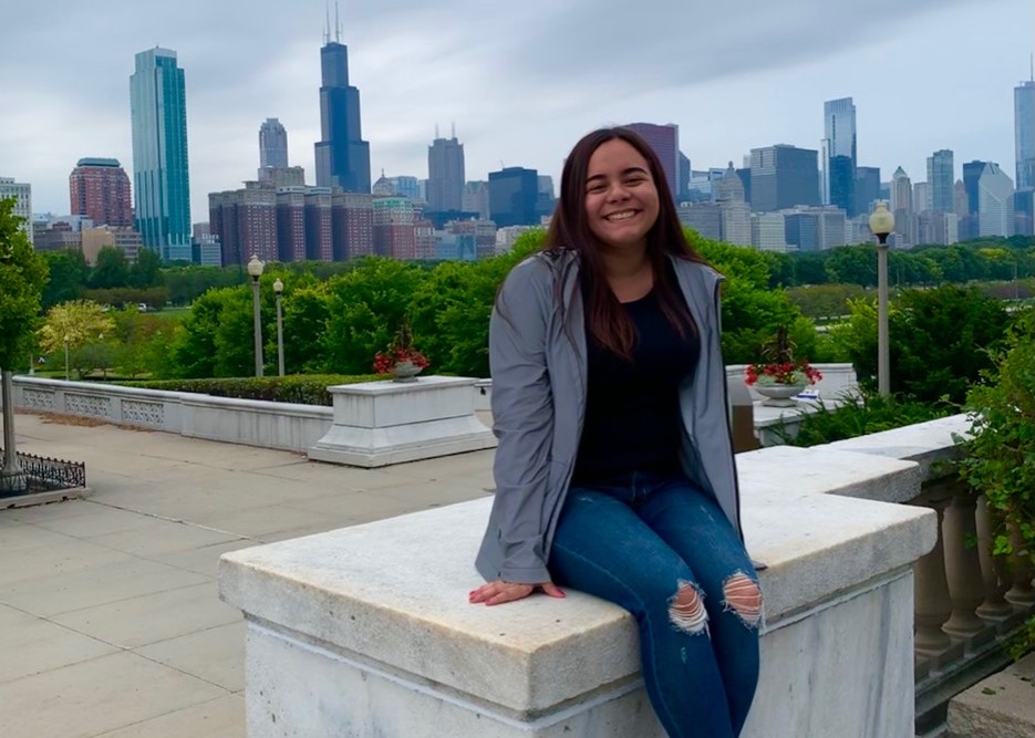 student Stephany Pichola sitting on a stone railing with the background of the Milwaukee skyline 