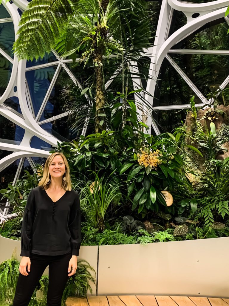 Rana Marks, wearing a black top and pants, stands against an architectural background of greenery.