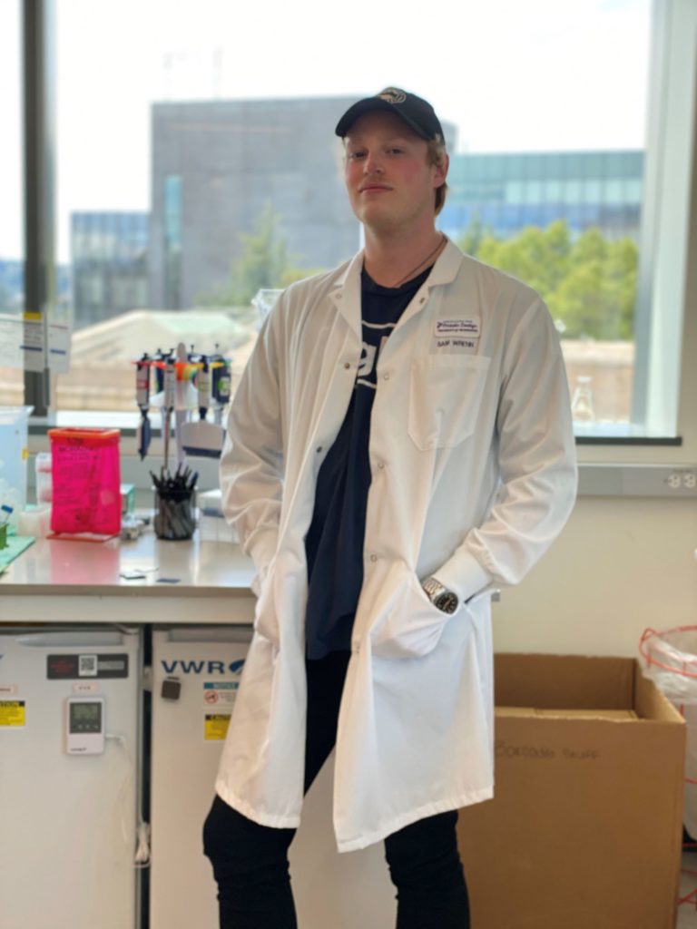 Sam Wrenn, wearing a baseball cap and a lab coat, stands in a laboratory.