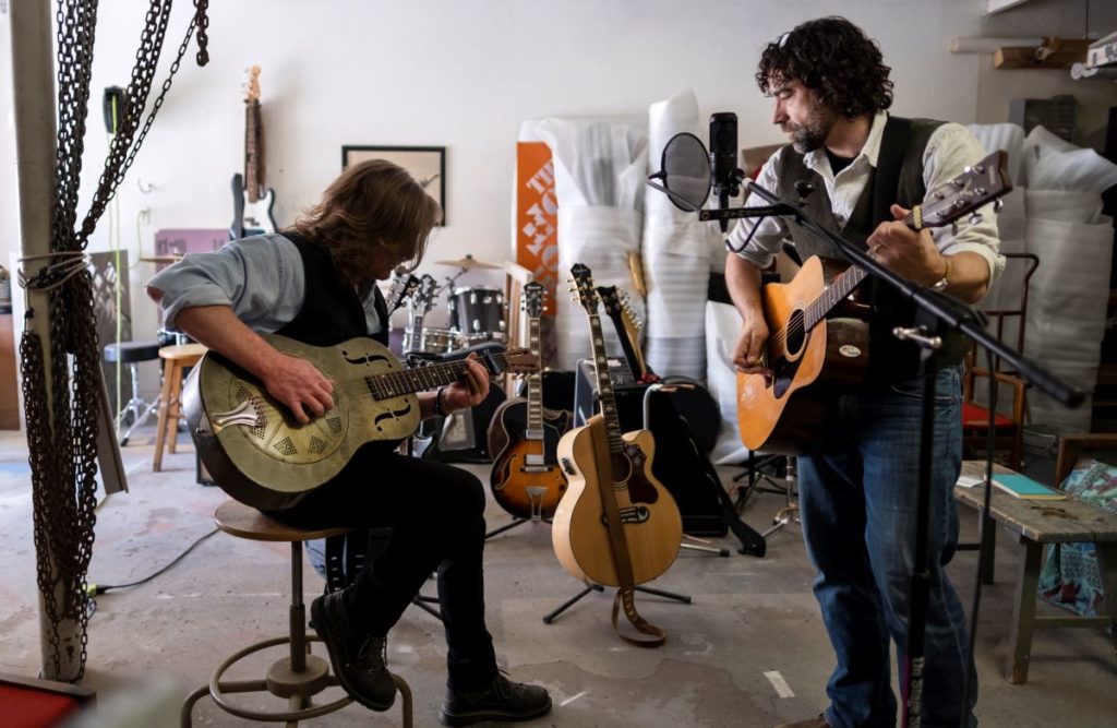 Rob Neilson sitting down playing the guitar and Jake Frederick standing next to him with a guitar