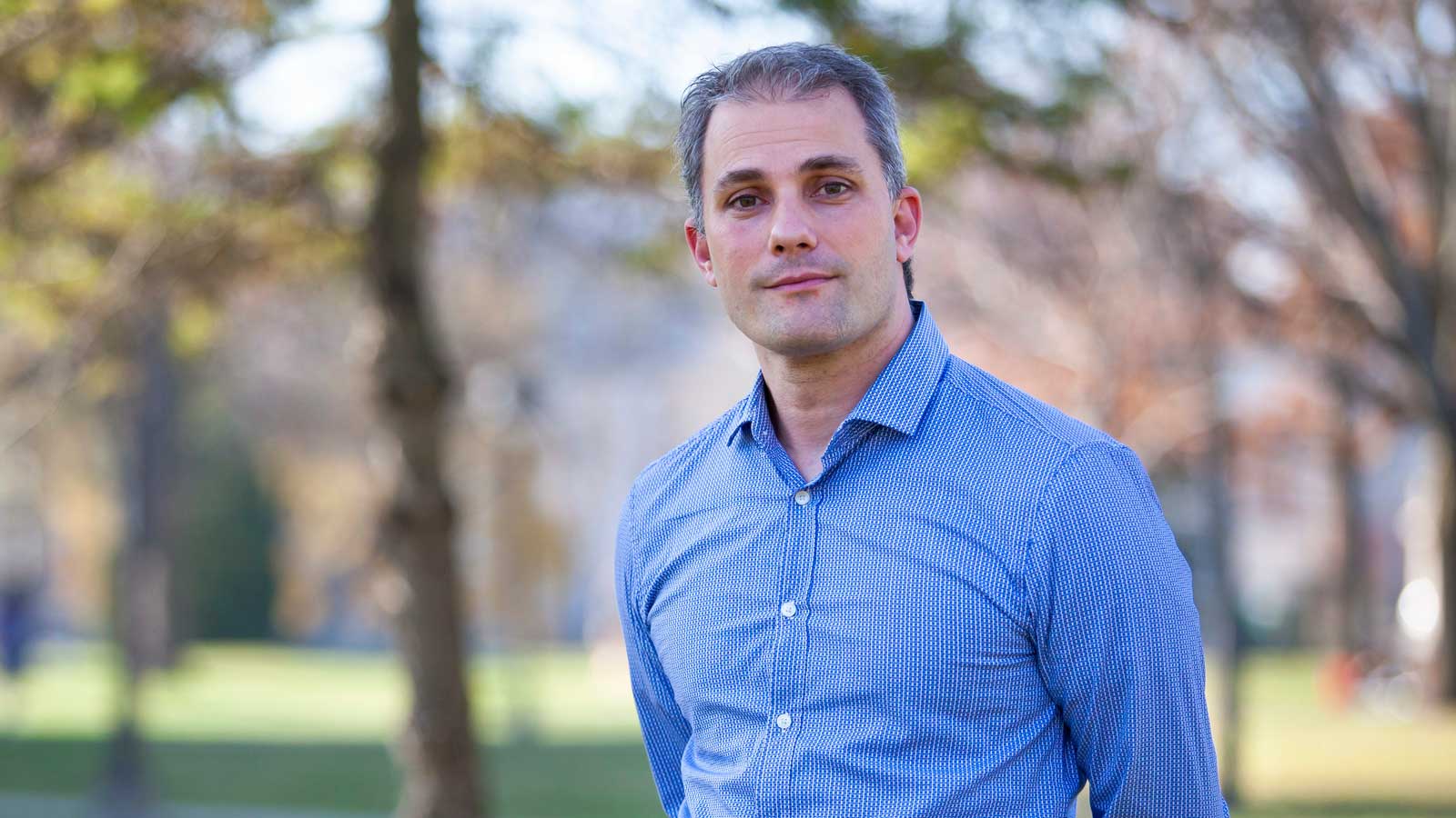 Scott Corry, wearing a blue button-up shirt, stands with his hands behind his back.