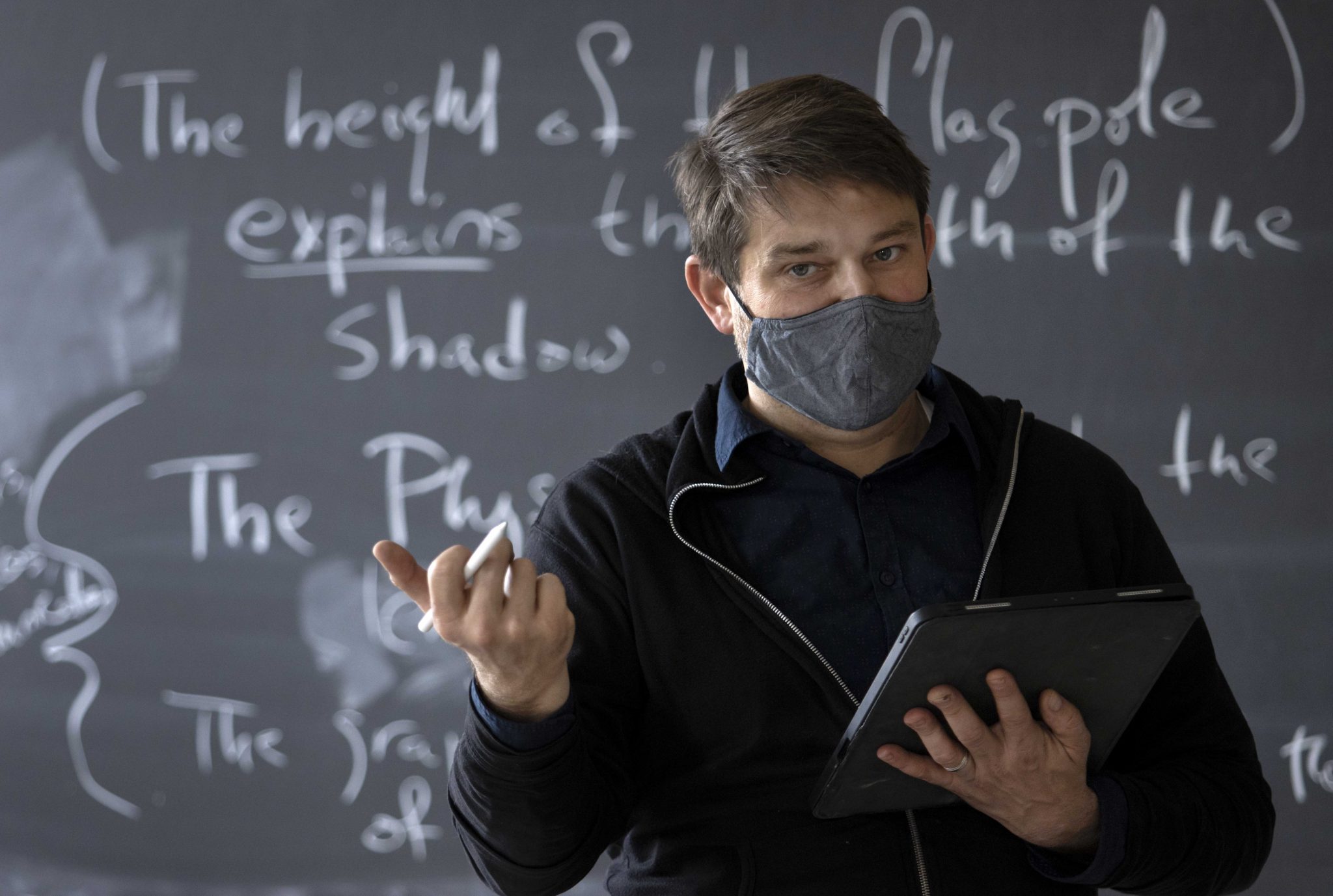 Mark Phelan, philosophy professor talks to class in front of blackboard, holding a tablet.