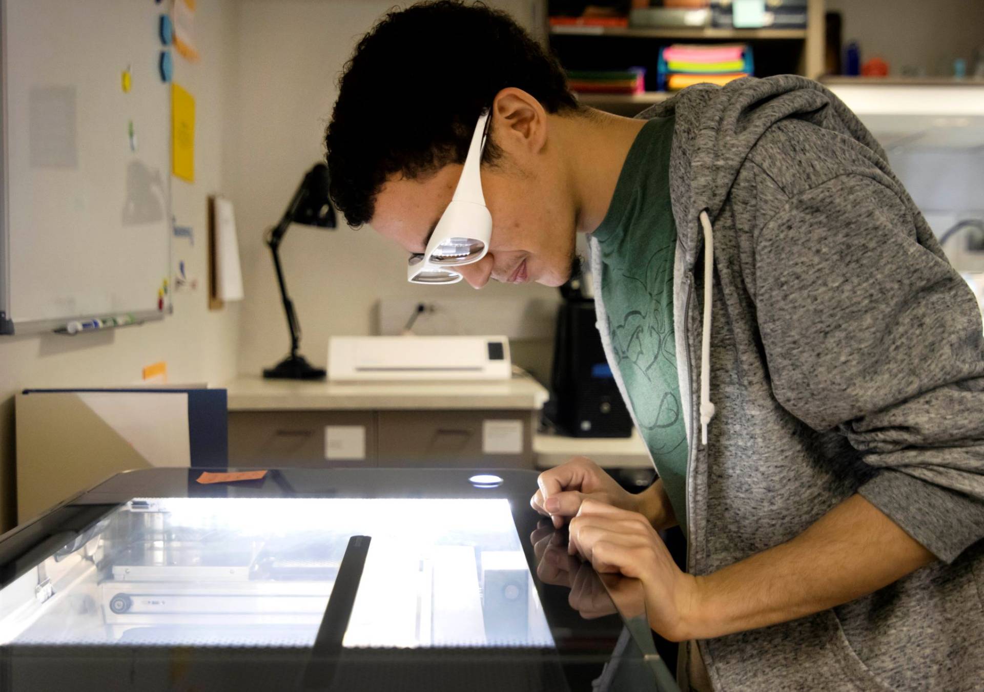 Student with protective eye gear leans over glass top to watch laser cutter.