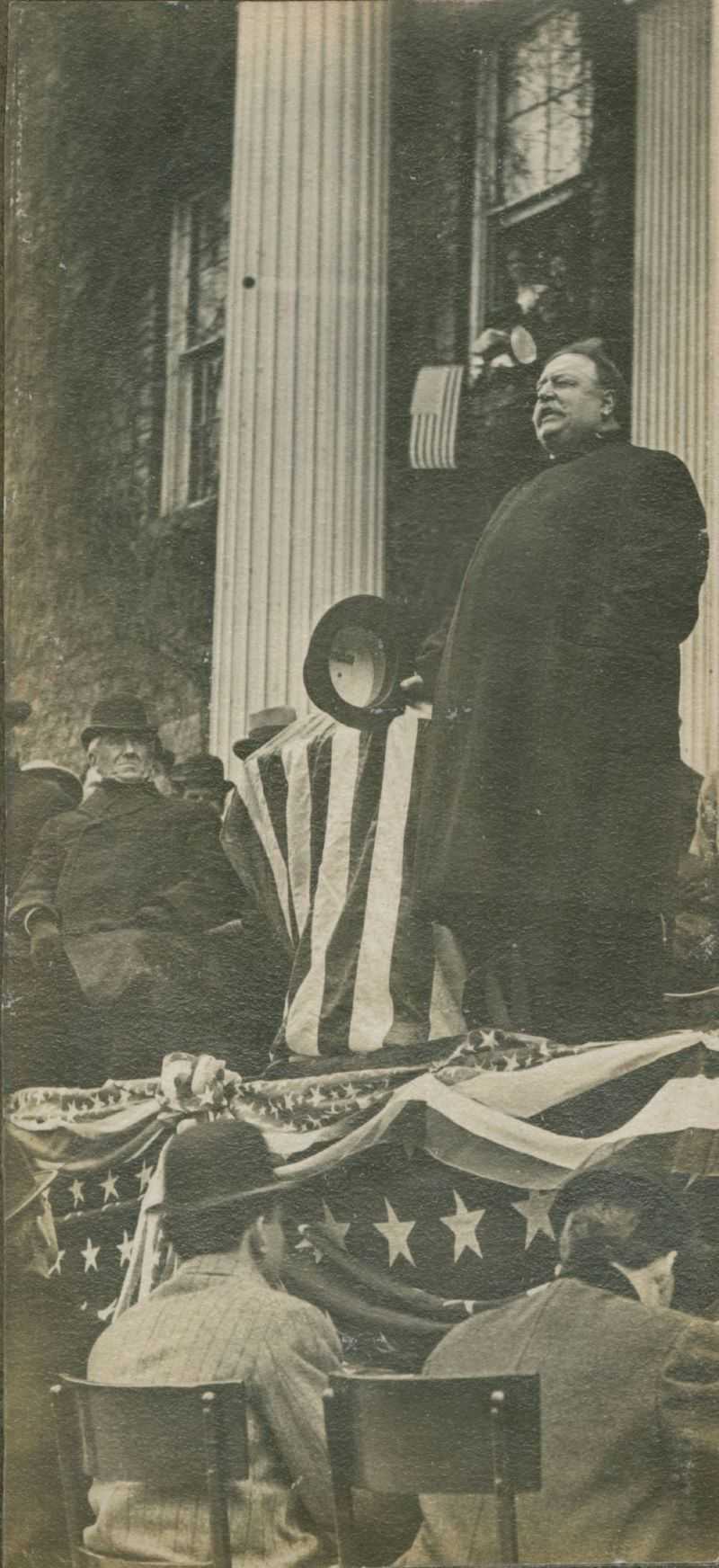 President Taft, wearing a long coat, holds his hat in his right hand as he stands on the steps of Main Hall while addressing a crowd on Main Hall Green.