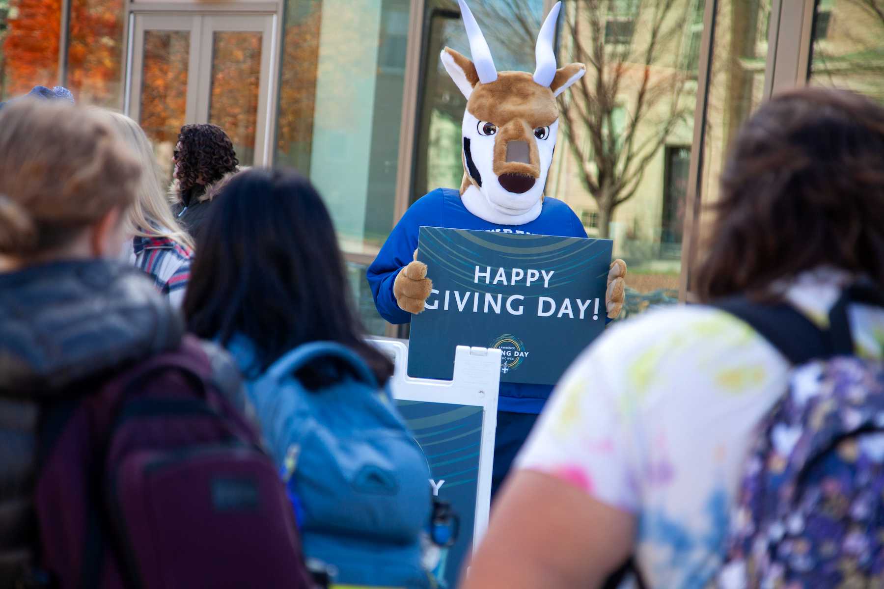 The antelope mascot holds a Happy Giving Day sign as it meets with students. 