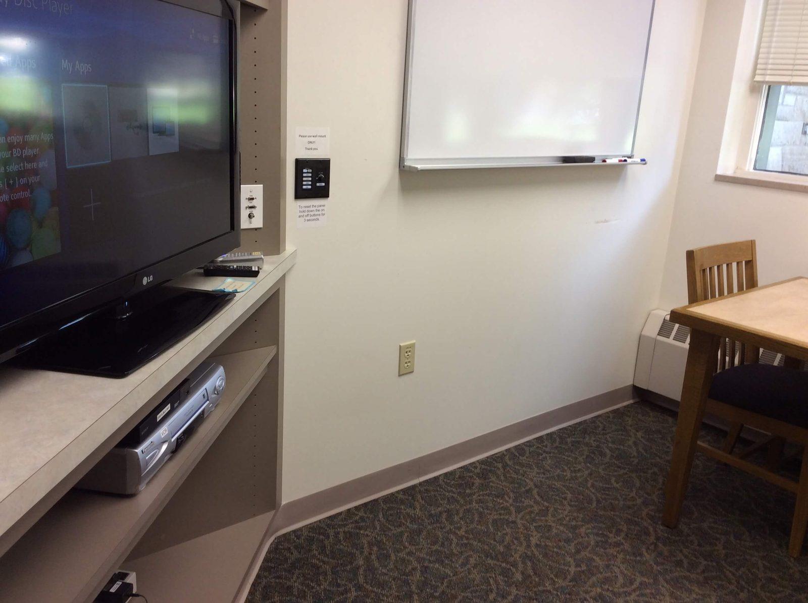 A library viewing room, which includes a dry erase board, table, two chairs, a large monitor, DVD/Blu-Ray player, and VCR