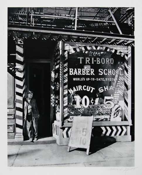 Black and white photo of a storefront, a striped column and doorway on the left and a display window with "Tri Boro Barber School" sign on the right