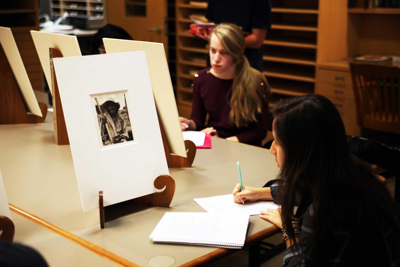 Two women sit studying artworks on a table in front of them, they take notes as they look.