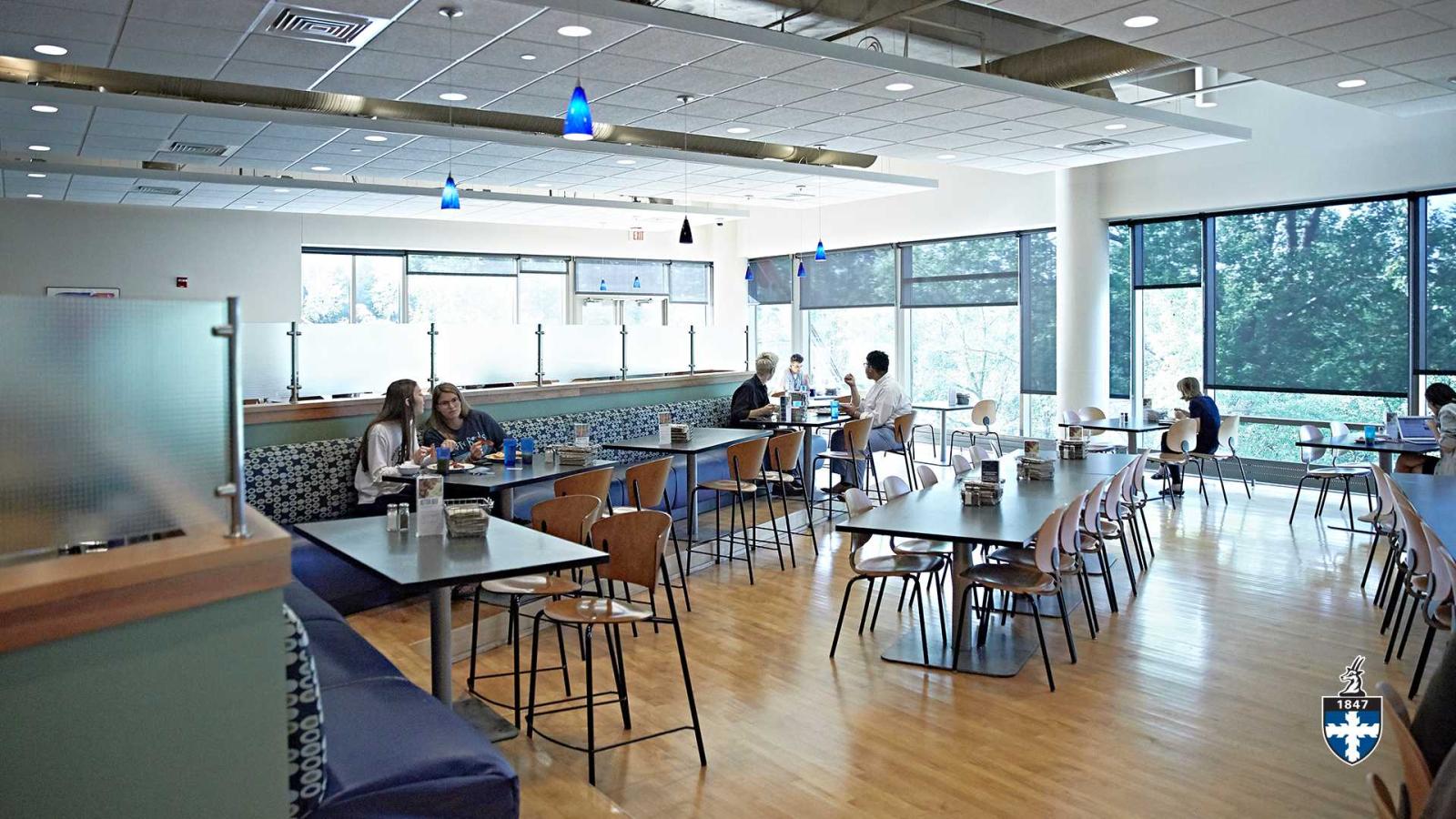 Students in open cafeteria eating with floor-length windows in background.