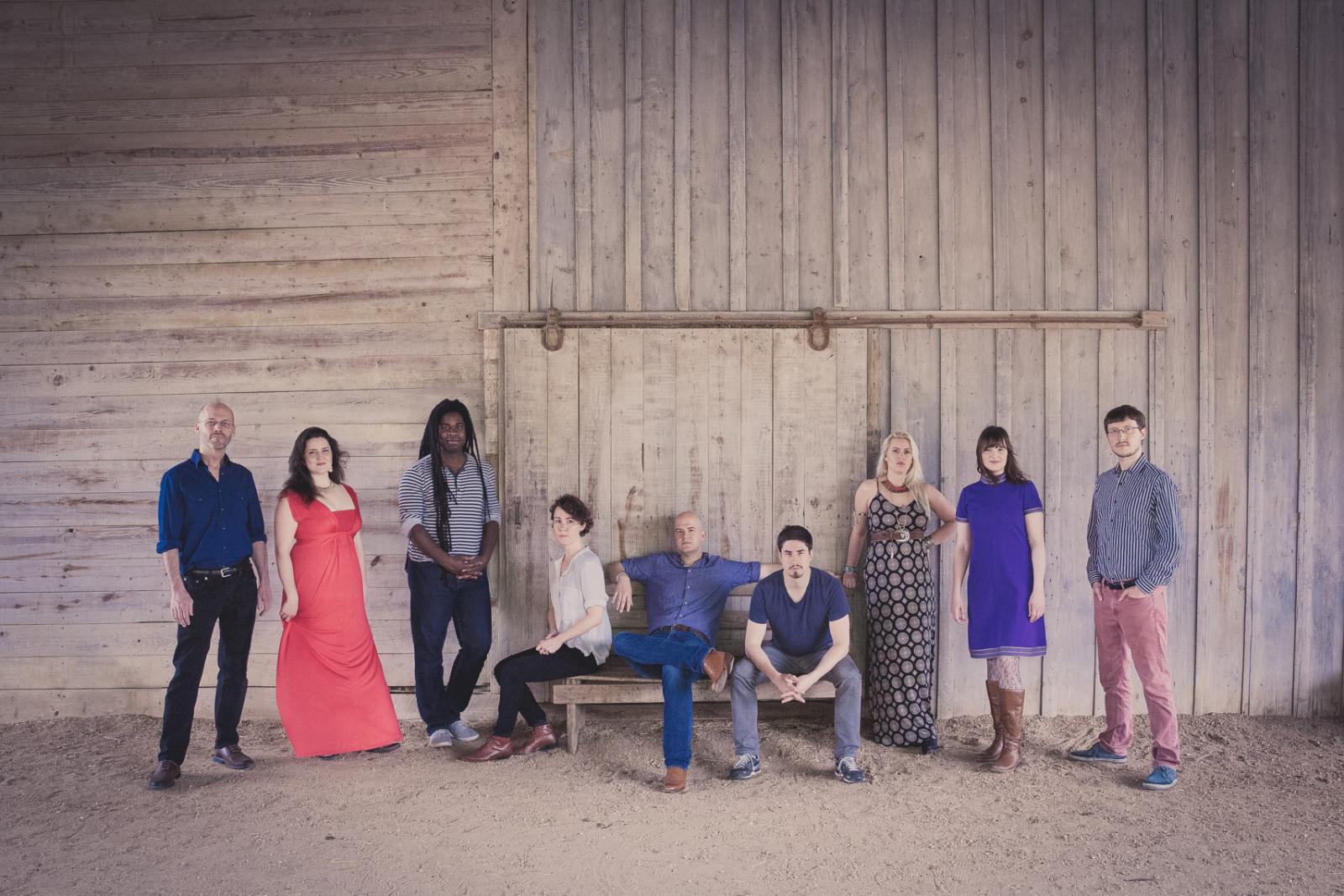 Nine members of the performing group Roomful of Teeth arranged in a line in front of a barn wall, center three performers seated. 