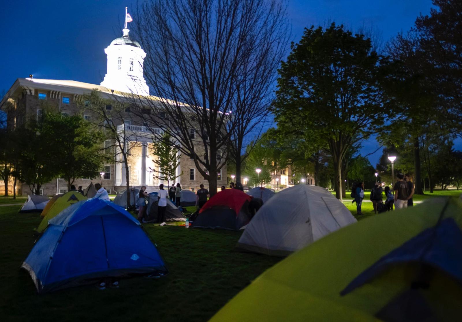 Colorful tents set up on Main Hall Green in front of a lit up Main Hall while students chat and help each other set up tents.