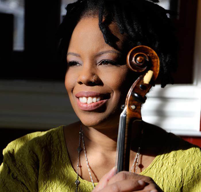 Violinist Regina Carter wearing a light green-yellow shirt, holding her violin and smiling. 