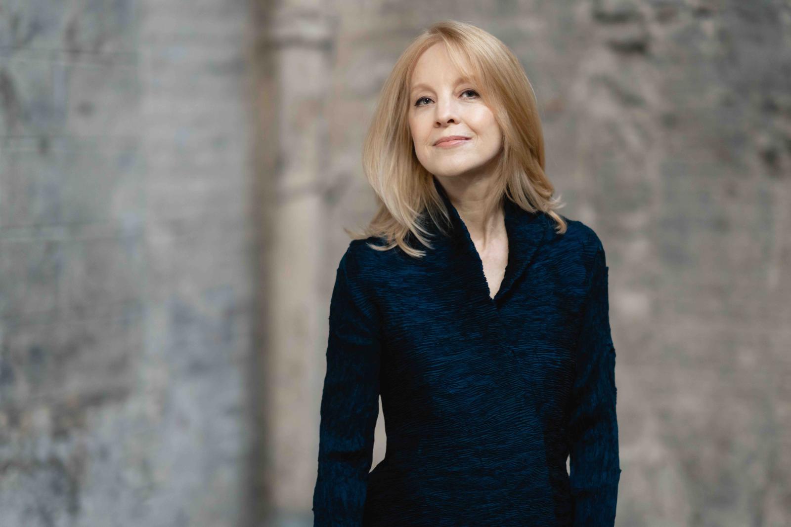 Musician Maria Schneider wearing a dark blue jacket, standing in front of a grey brick wall which is blurred. 