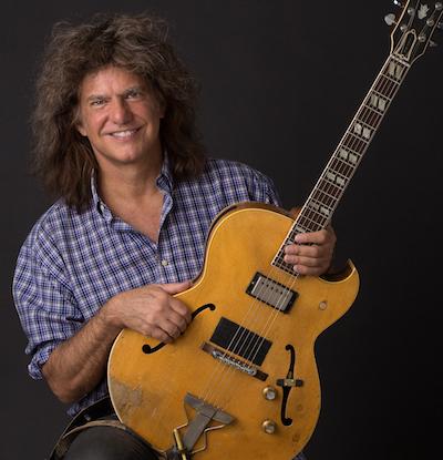 Musician Pat Metheny wearing a blue checked button up shirt and holding his guitar in front of a dark grey background.