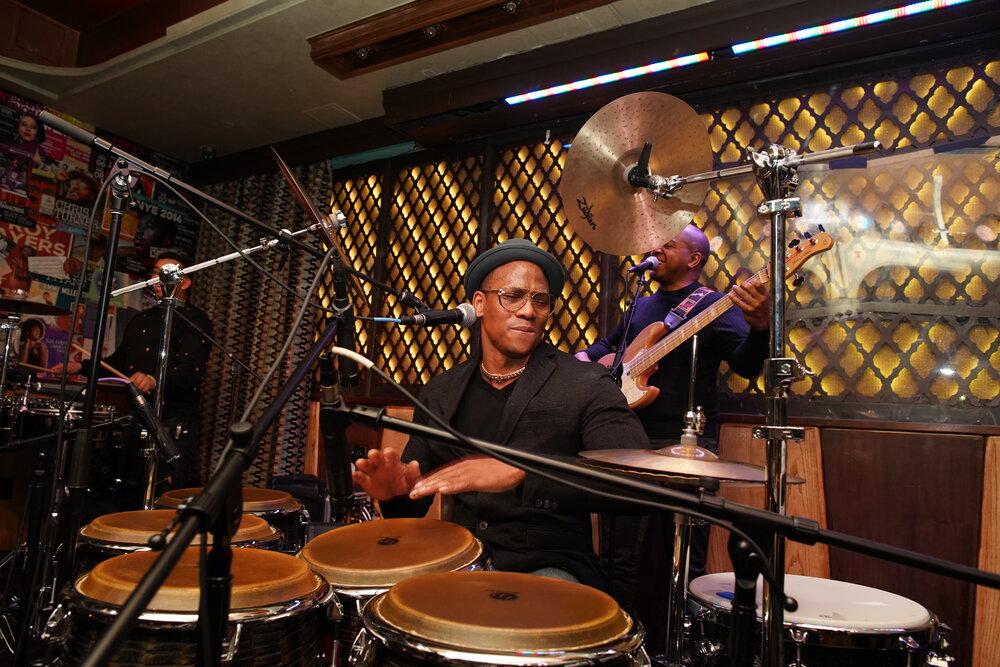 Musician Pedrito Martinez playing the drums, another musician playing guitar behind him. 