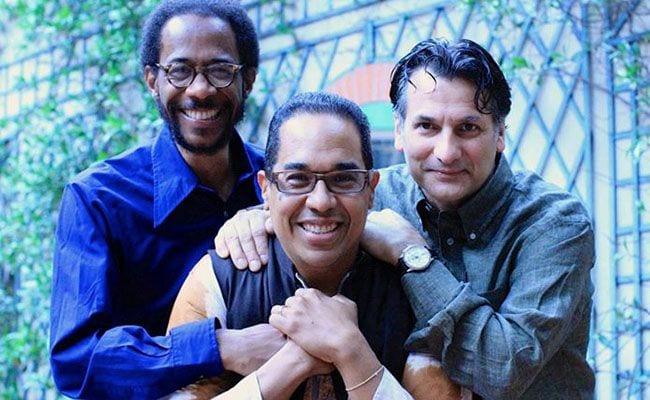 Musicians Danilo Perez, John Patitucci and Brian blade grouped together and smiling. Perez in front with Blade's arm around him and Patitucci's hands on his shoulders.