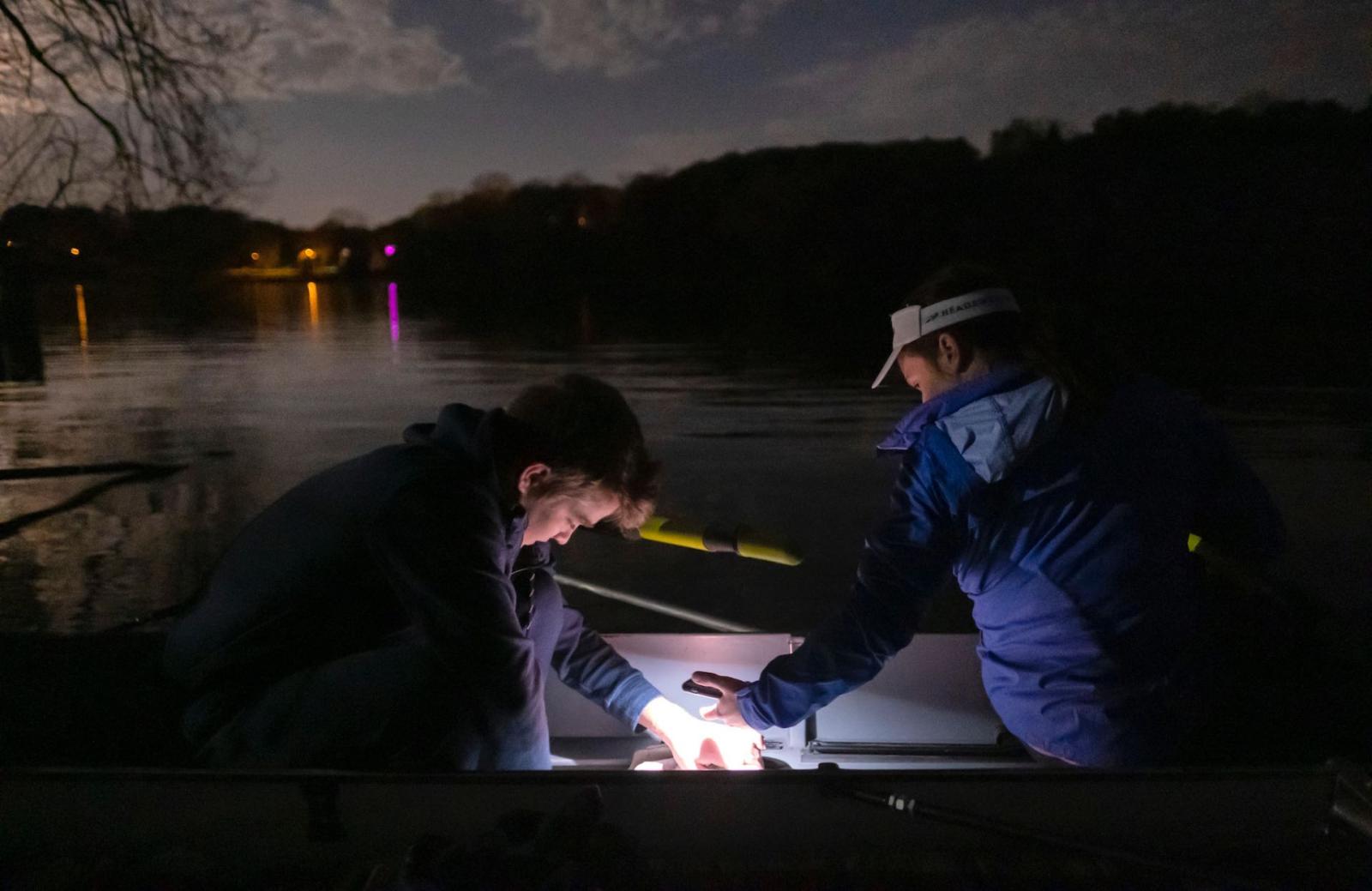 Two students crouching in the dark. One holds a light as the other prepares their boat by the river as the sun begins to rise.