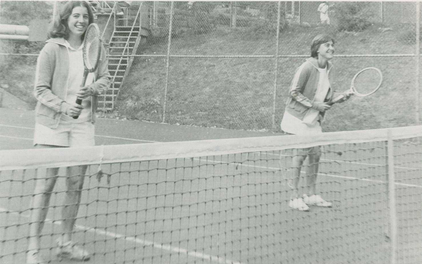 Two members of the women's tennis team stand on a tennis court behind a net
