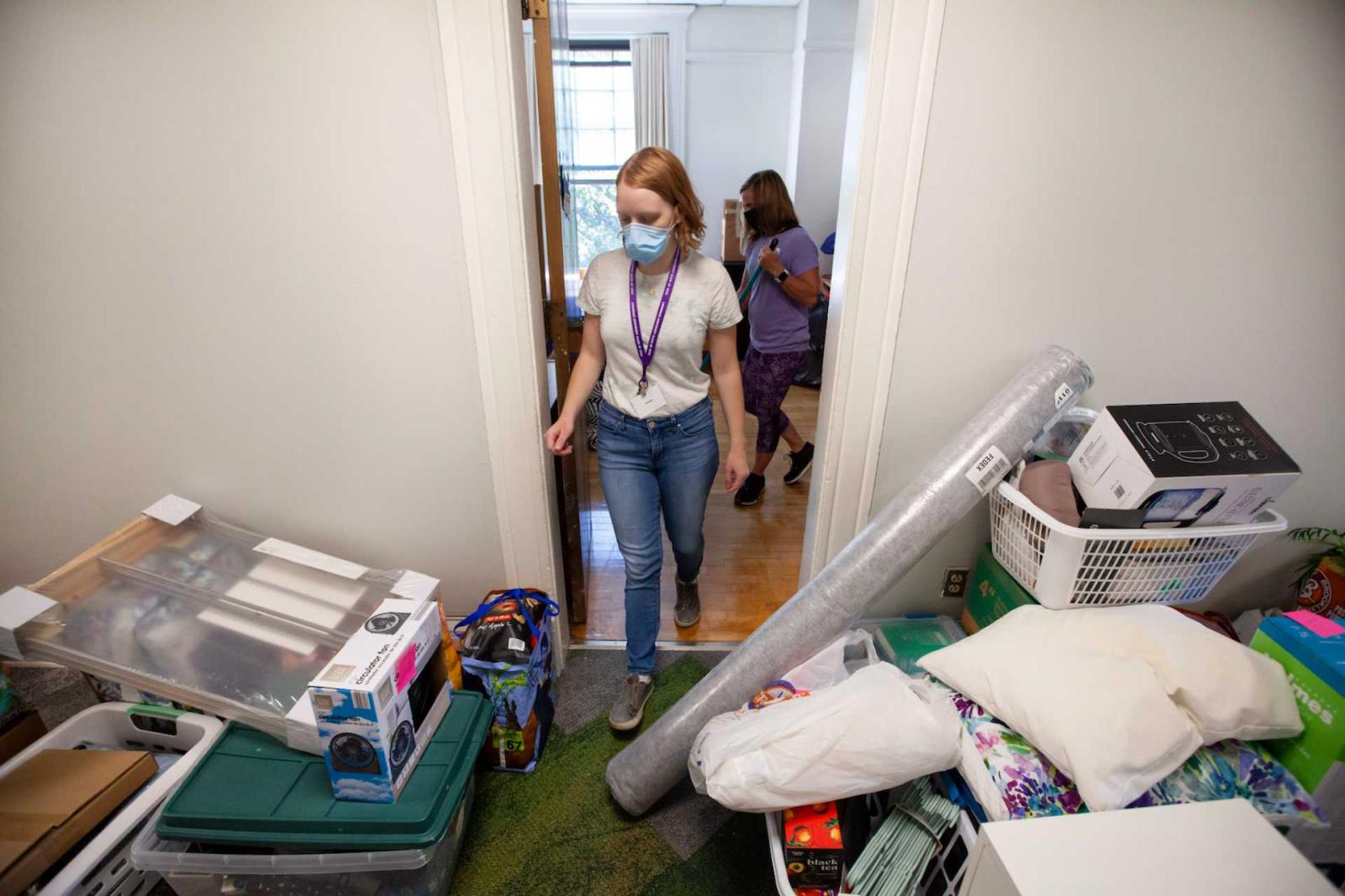 Student walks into dorm room filled with unpacked furniture and luggage.