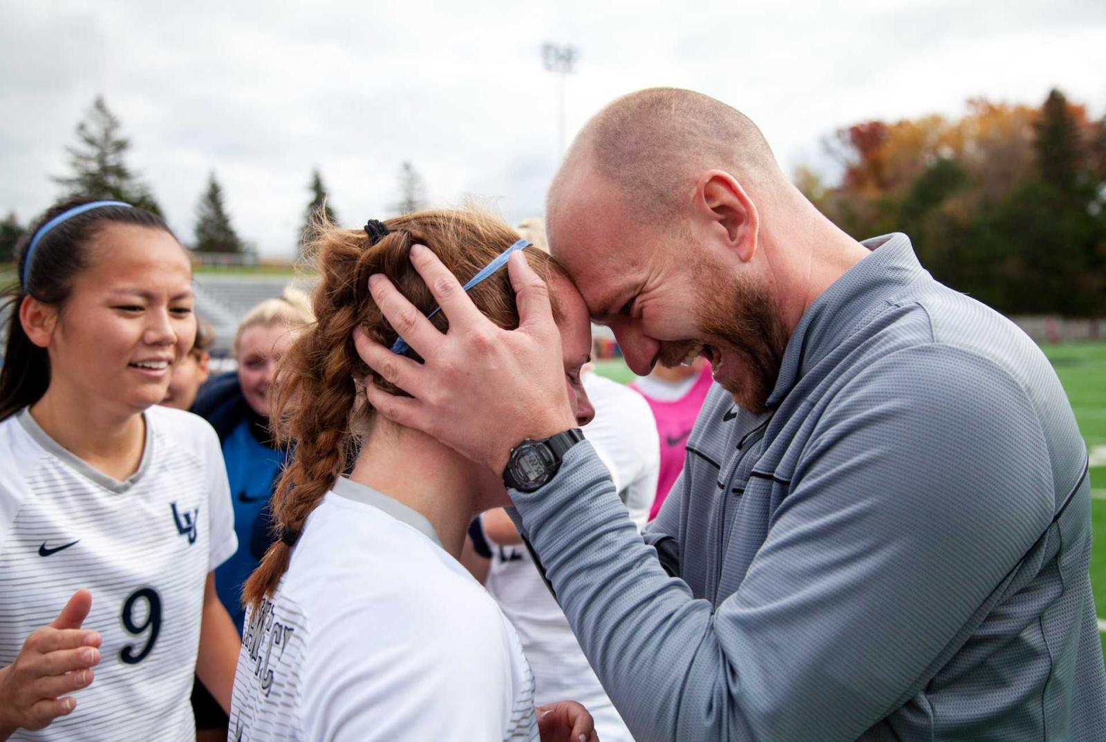 Coach Joe Sagar presses his forehead to Emma Vasconez's forehead as teammates around them cheer.