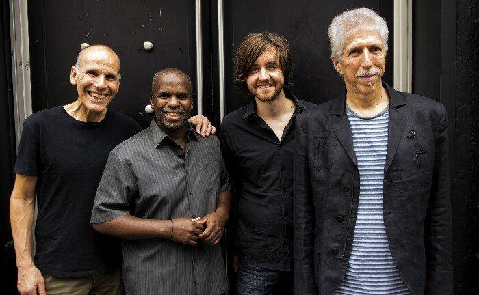 The musicians of the performing group The Yellowjackets, standing in a row in front of a large window. It is night so the window is dark.
