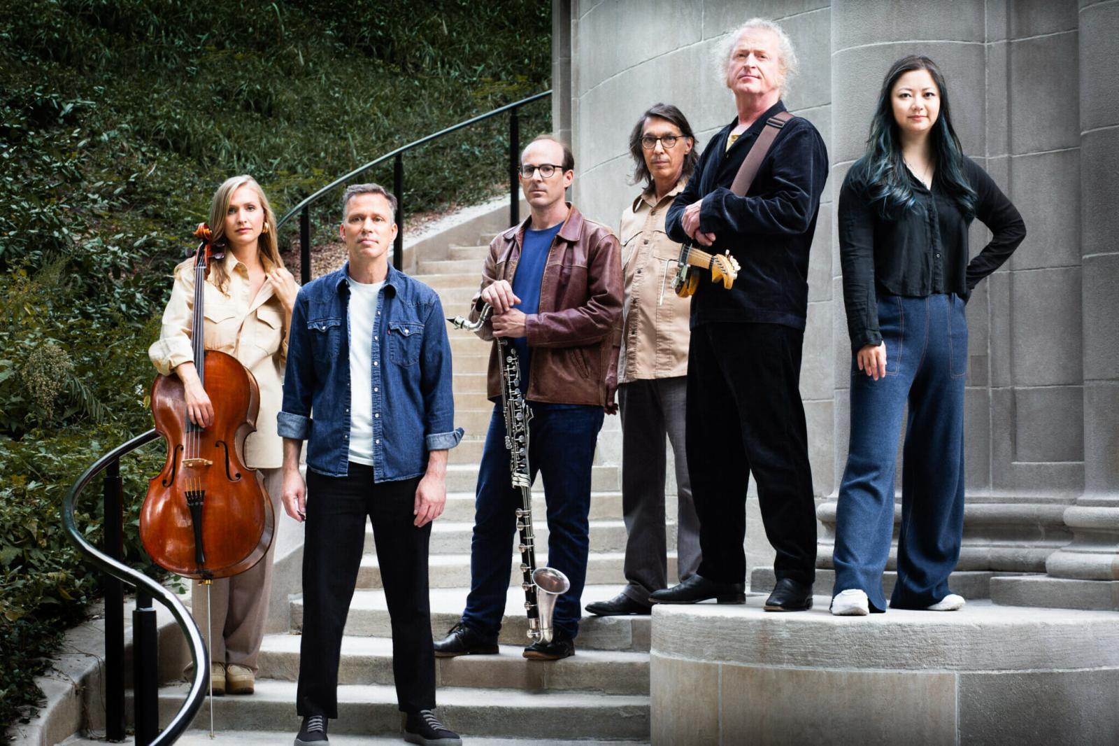 The six performers of Bang on a Can All Stars stand across a set of steps with a railing running down the left side. The Cellist and the Bass Clarinetist have their instruments. 