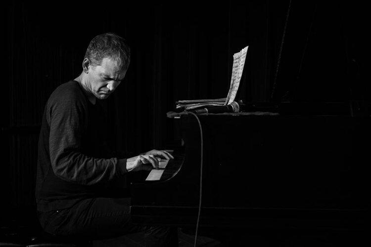 Black and white image of Brad Mehldau seated at a piano looking to his right. 