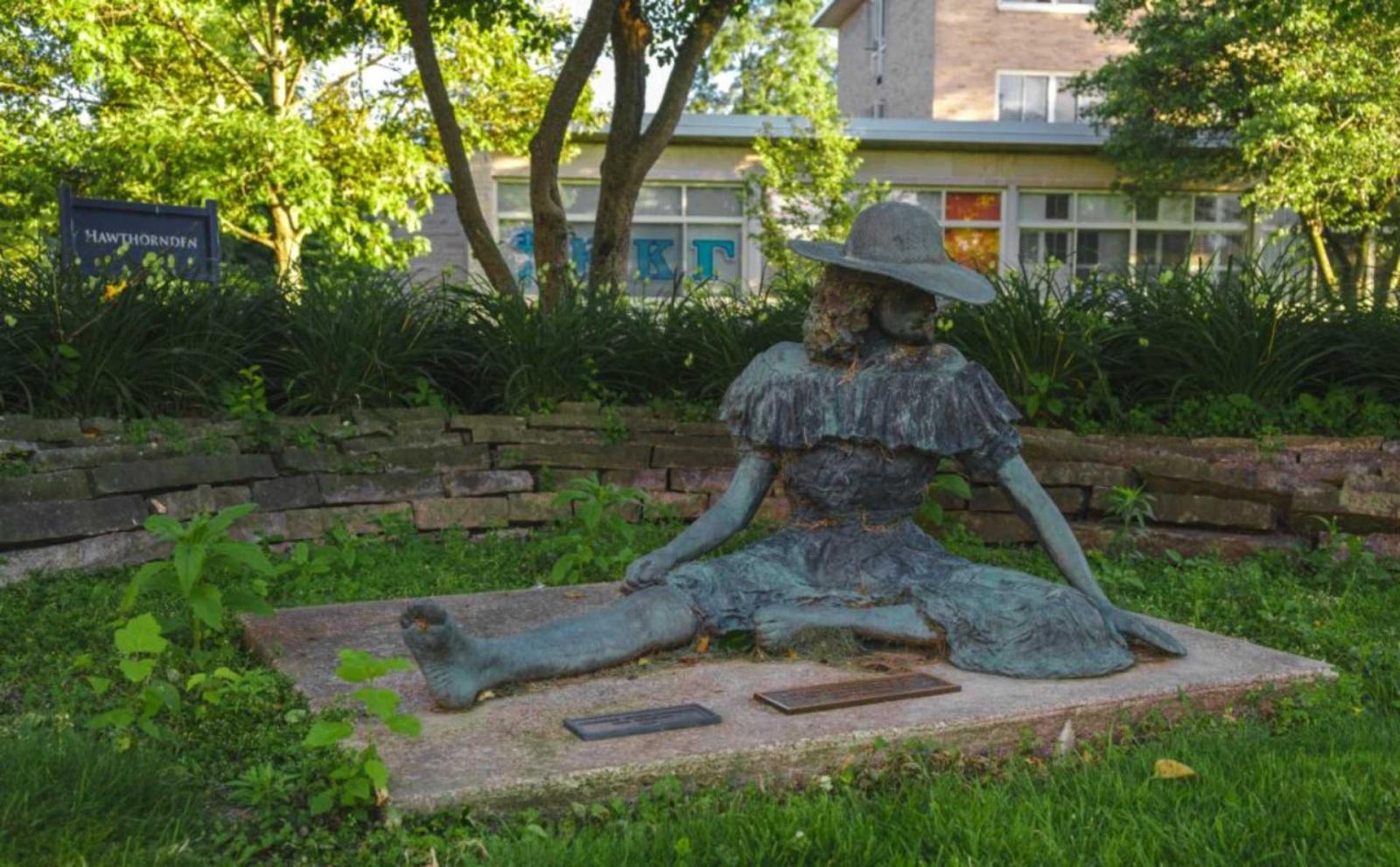 A patinaed metal sculpture of a girl in a dress and hat sitting on the ground