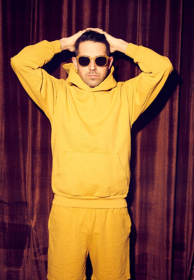 Drummer Mark Guiliana wearing a yellow track suit standing in front of a red velvet curtain wearing sunglassess with his hands on his head.