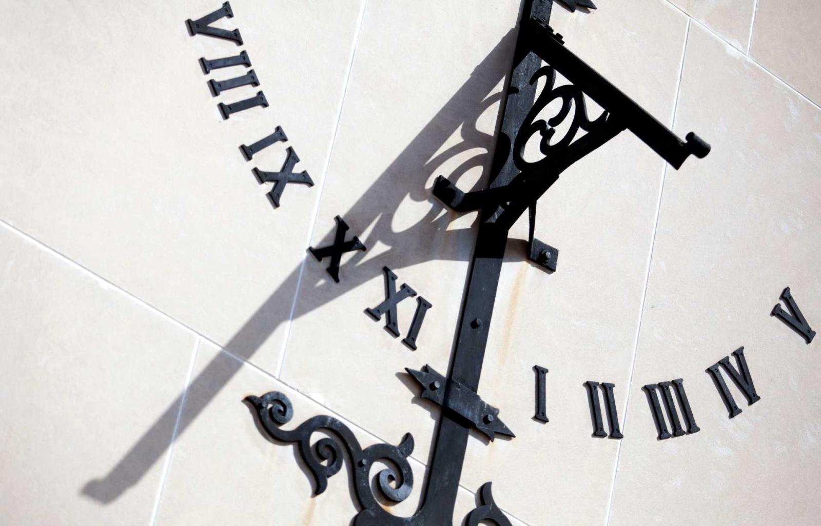 Black Roman numerals arranged in a half circle on a stone wall; an ornate arm intersects the half circle in the middle and casts a shadow onto the numbers. 