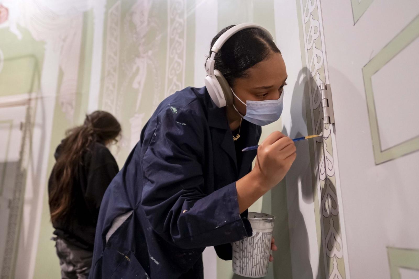 Student wearing headphones and painter's coveralls paints details onto a doorframe while holding a small cup of paint.