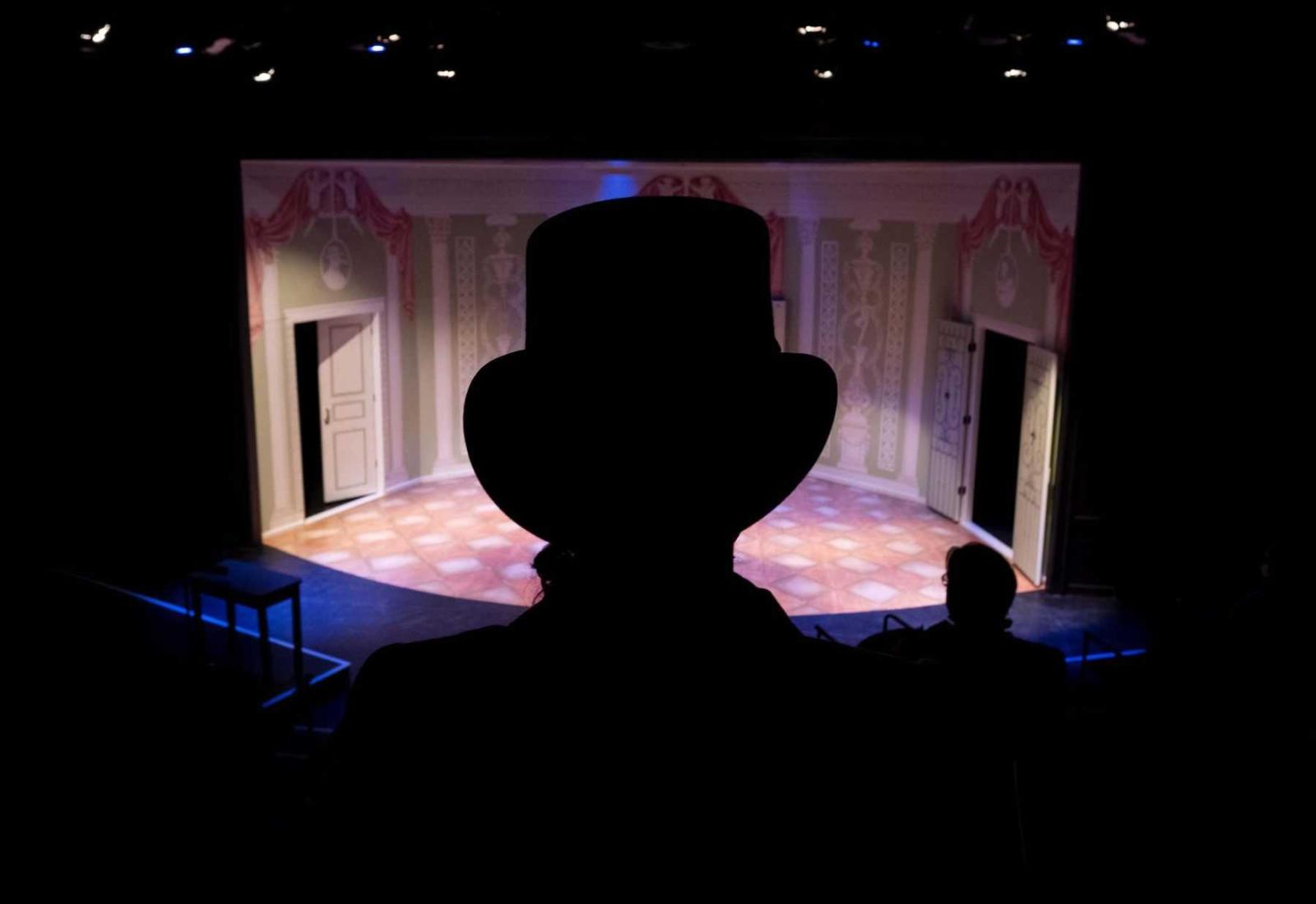 A cast member in costume is silhouetted from behind against the stage while watching rehearsal.