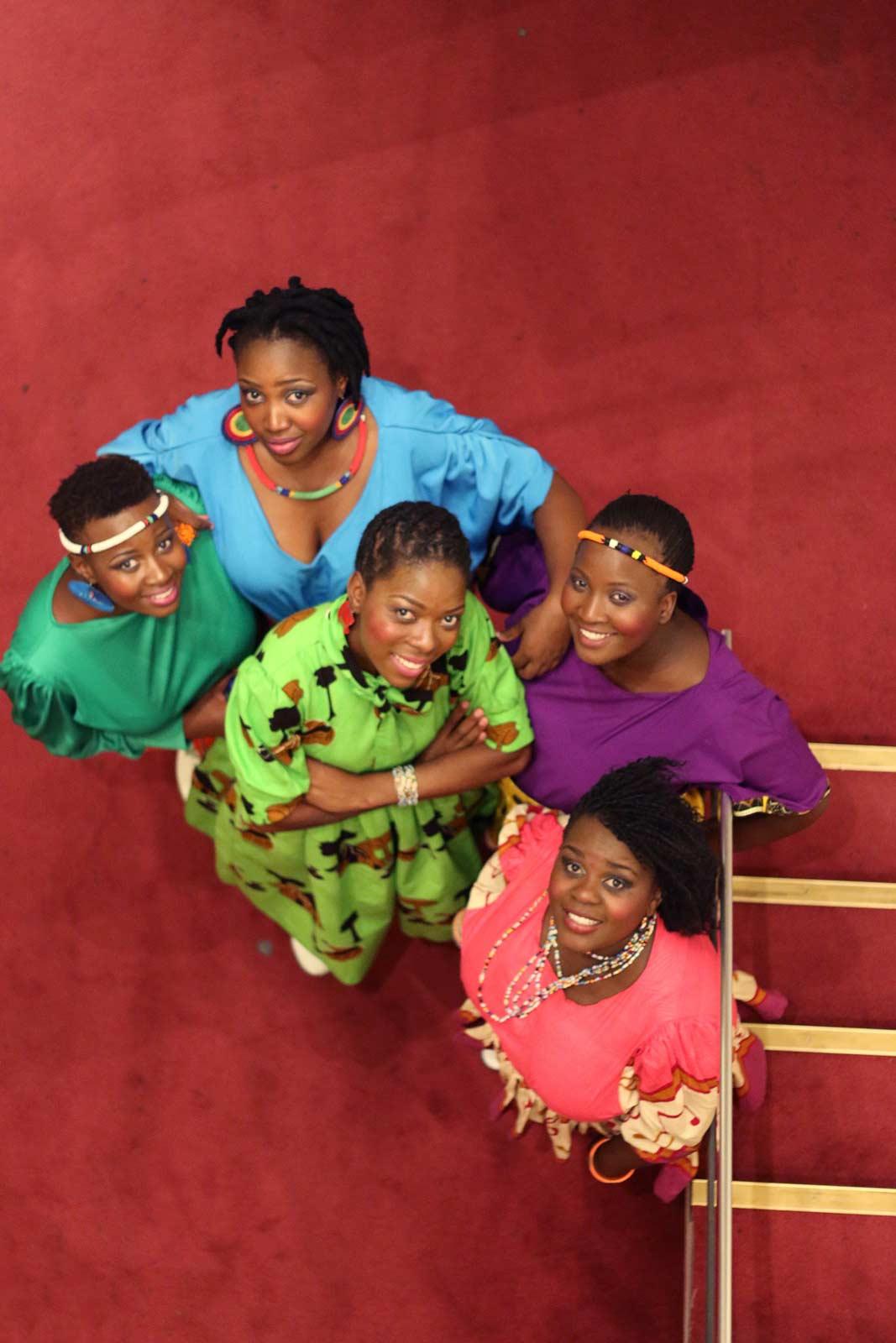 Performers of Nobuntu wearing colorful dresses and looking up at the camera standing on red carpet.