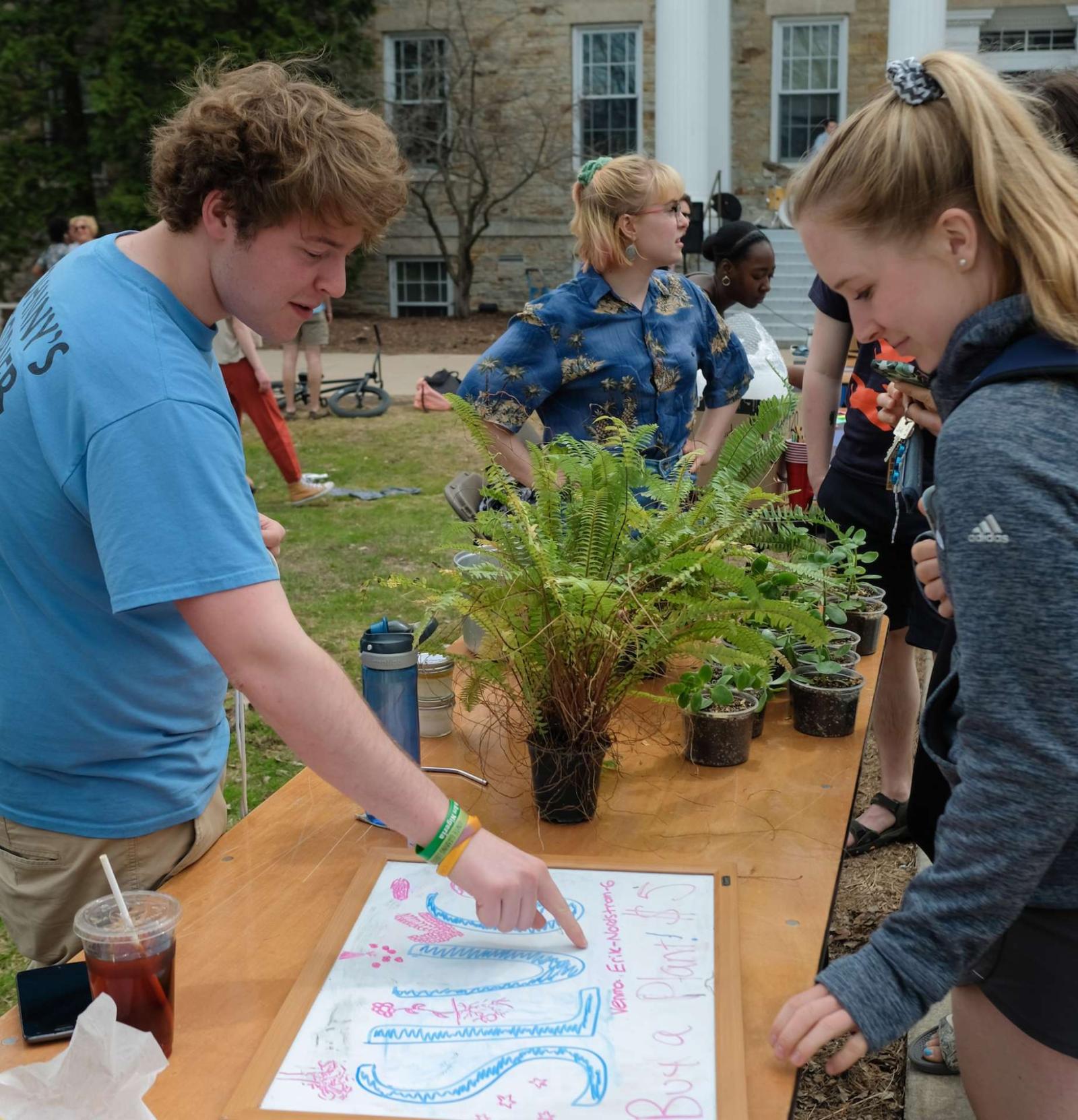 Students stand around table with ferns and other small pots of plants. A sign between two students say "SLUG."