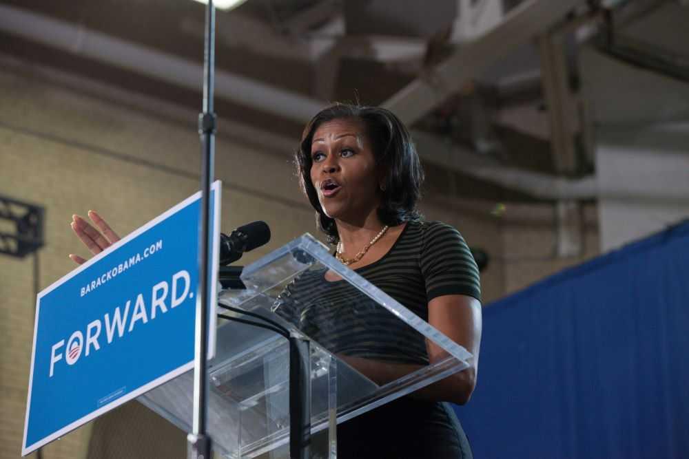 Michelle Obama speaks from a stage in Alexander Gym. A sign reading FORWARD is on front of podium.