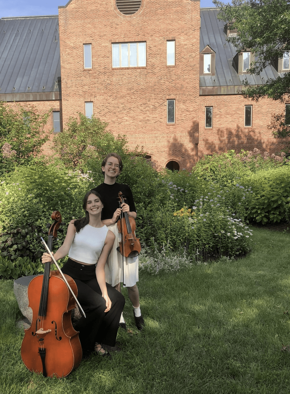Two students hold a violin and cello in a garden.