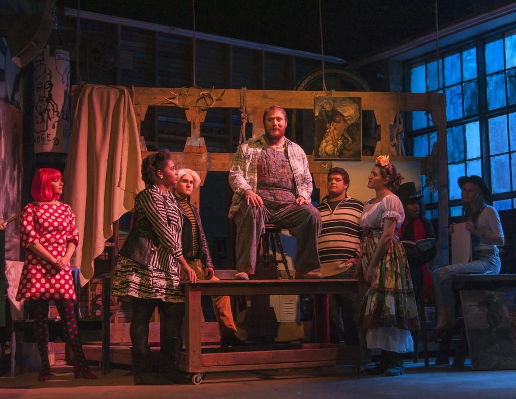 Production photo of the musical, Godspell: man sitting on chair on top of table while 6 others stand around the table and watch him.