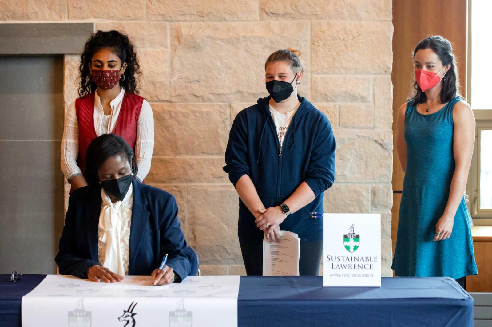 President Laurie Carter signs the Climate Commitment at a table while Adya Kadambari, Em Gajewski and Relena Ribbons stand behind her.