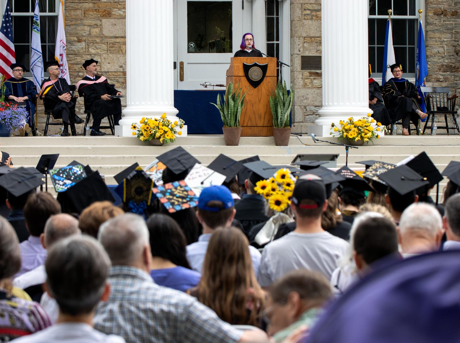 Helen Kramer stands at a podium on the steps of Main Hall while an audience of capped graduates watches.
