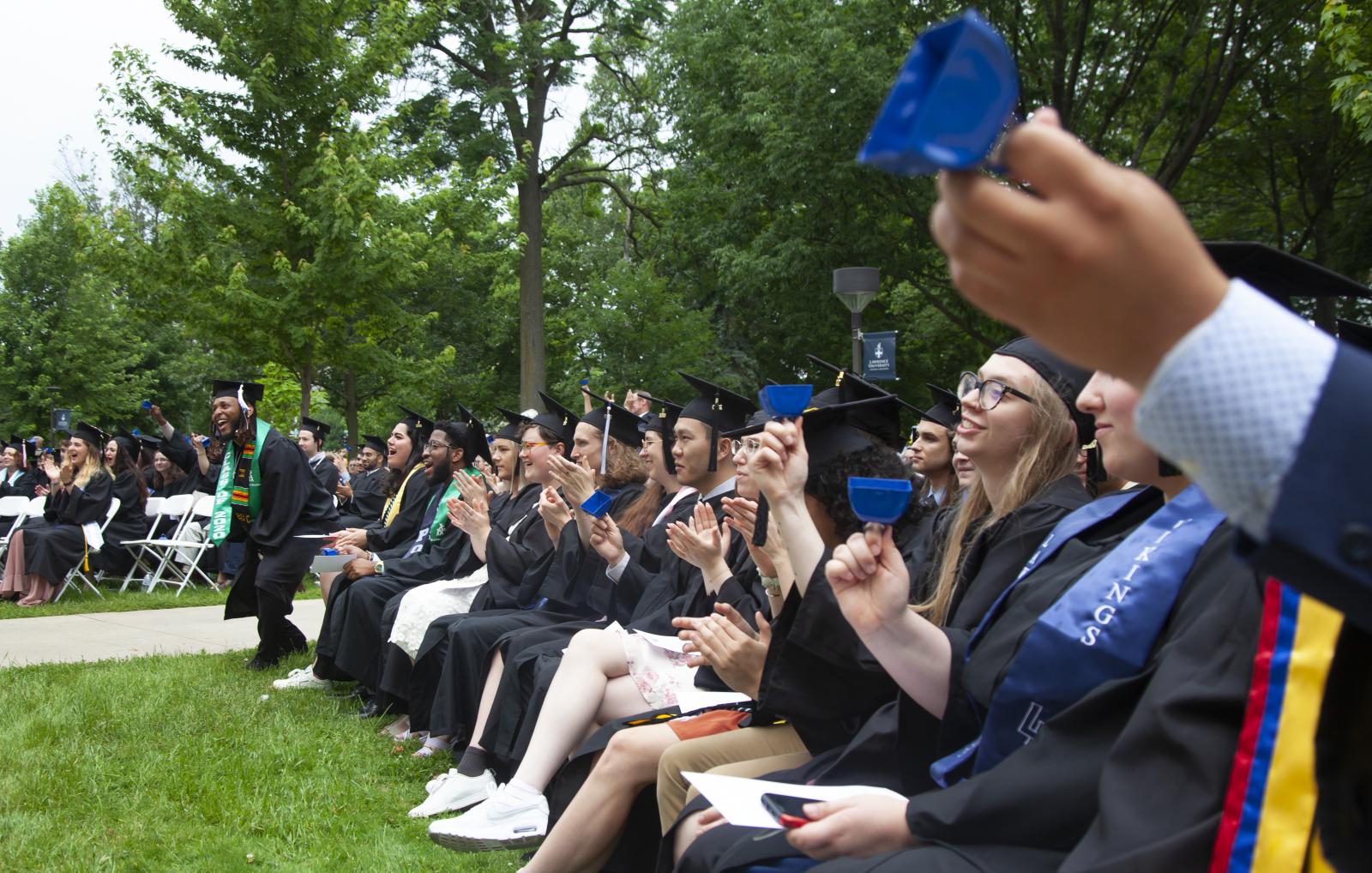 Graduates dressed in cap and gown cheer, applaud, and ring cowbells. 